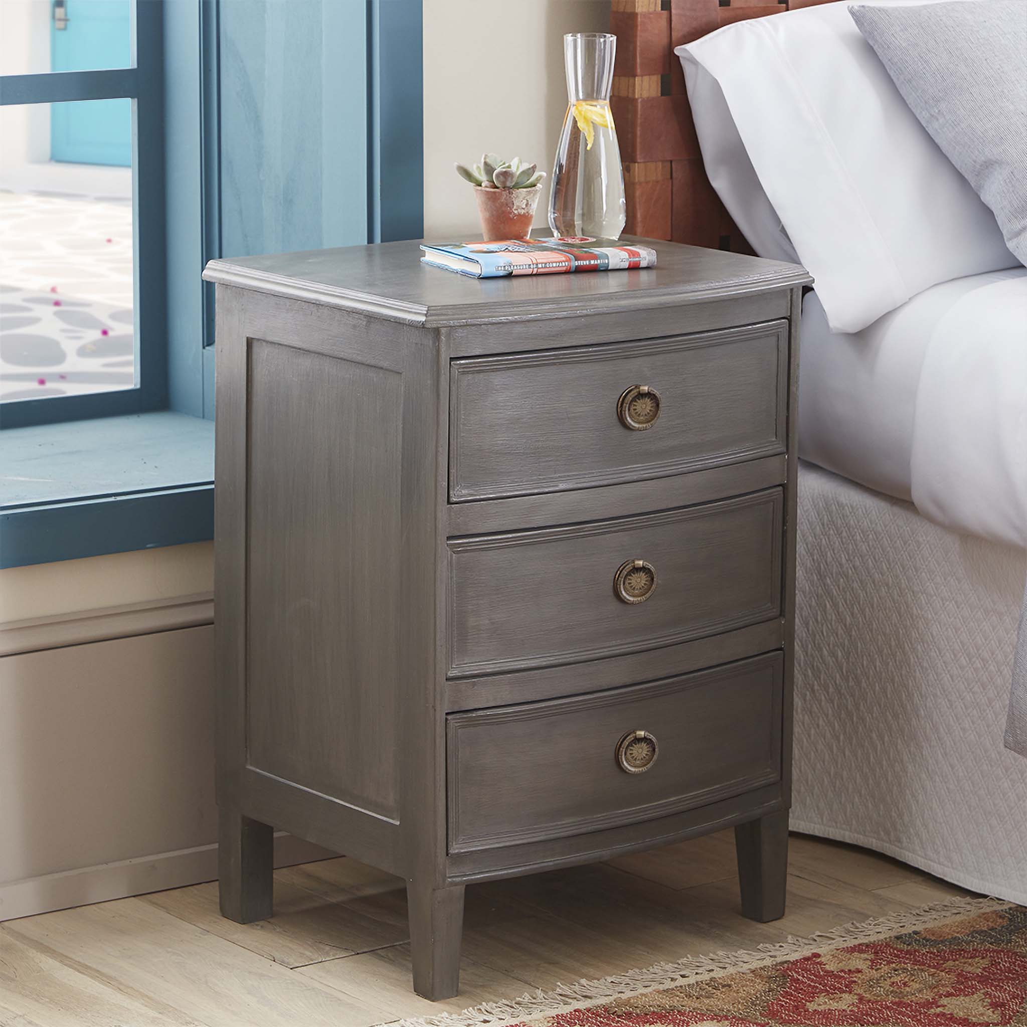 Kunj Three Drawer Chest – Pumice finish, styled as nightstand with books and glassware, bedroom setting.